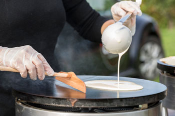 Gros plan sur les mains de Suzette tenant une louche et un rozell et s'appretant à tourner la pâte à crêpes sur la crêpière